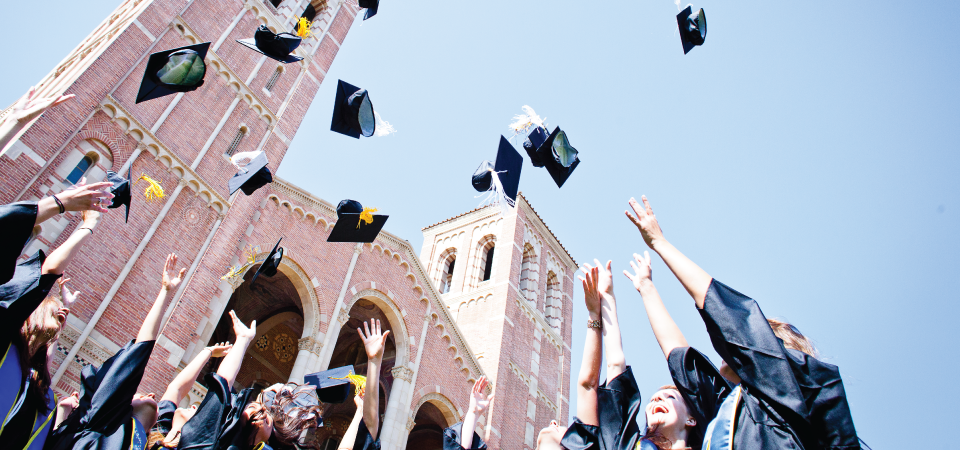 Students in commencement attire can be seen throwing their graduation caps into the air in front of a UCLA campus building.