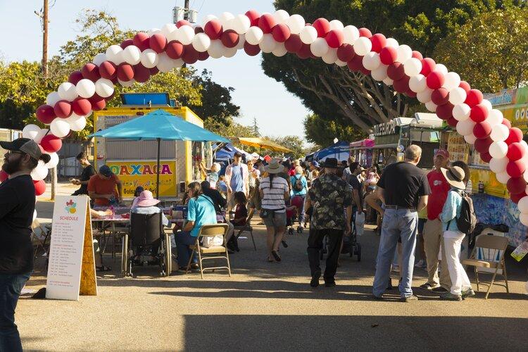 The photo shows a stretch of the Westchester Arts and Music Block Party. There are many pedestrians walking through the area, and a red and white balloon arch signifies the entrance to vendors and stands at the block party.