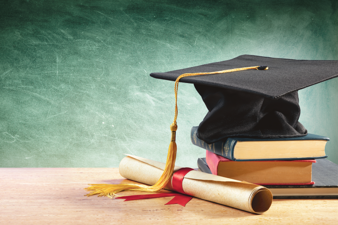 image of a black graduation cap on a stack of books beside a rolled graduation certificate wrapped neatly in a red ribbon. All items are sitting on a wooden desk in front of a chalkboard.
