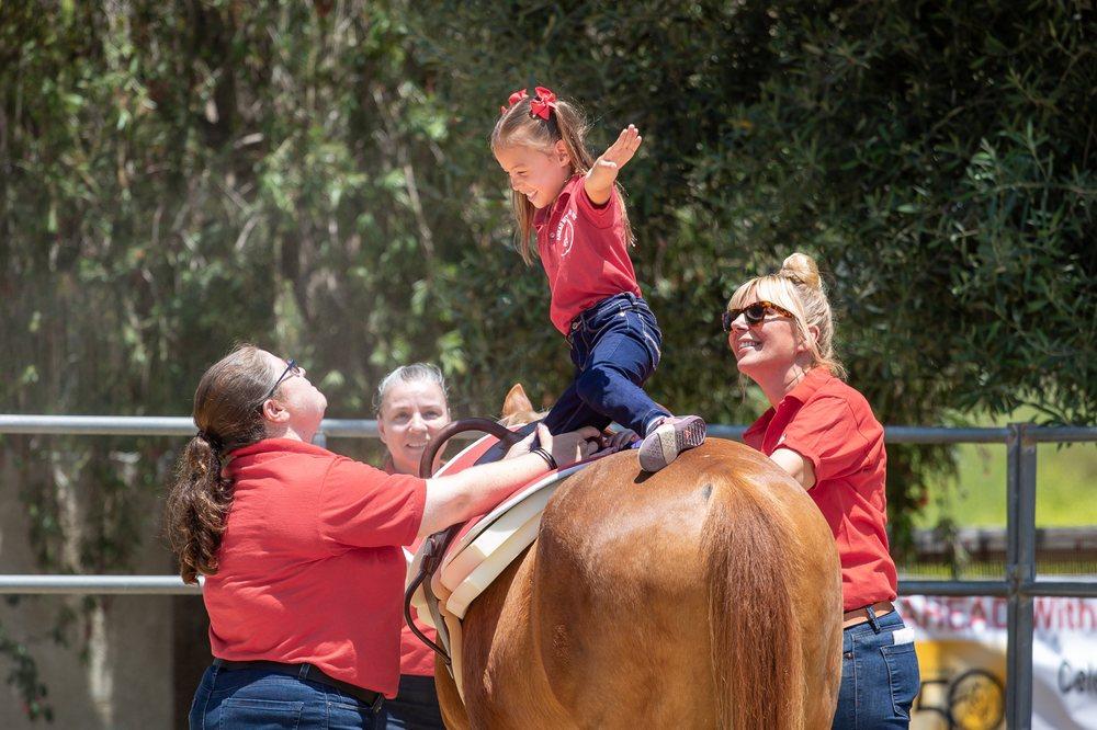 Image displays a happy child standing while riding a horse with the assistance of staff from AHEAD with horses.