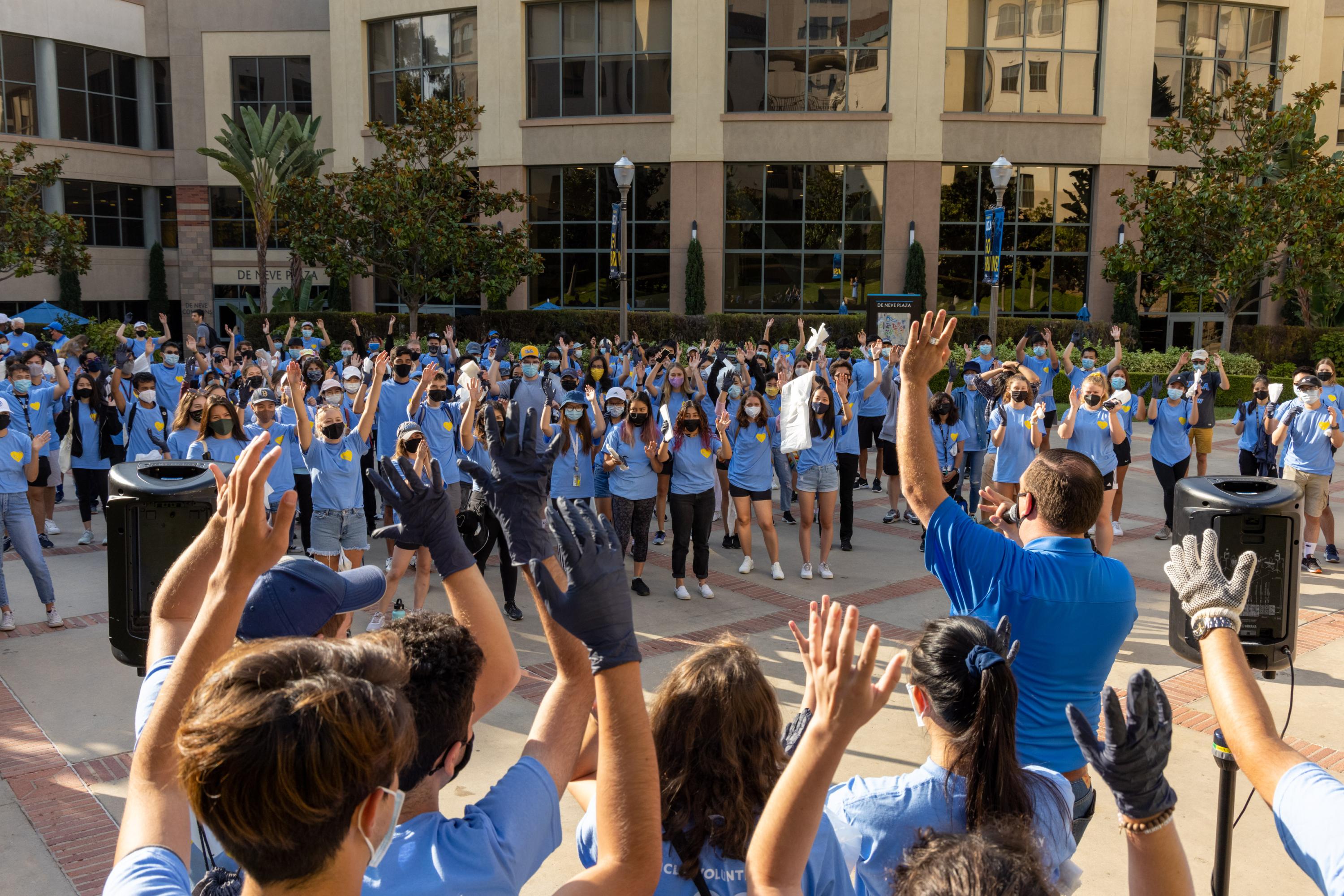 A large group in blue shirts gathers outdoors, raising hands in a volunteer or community event. A speaker leads them in front of a modern building.