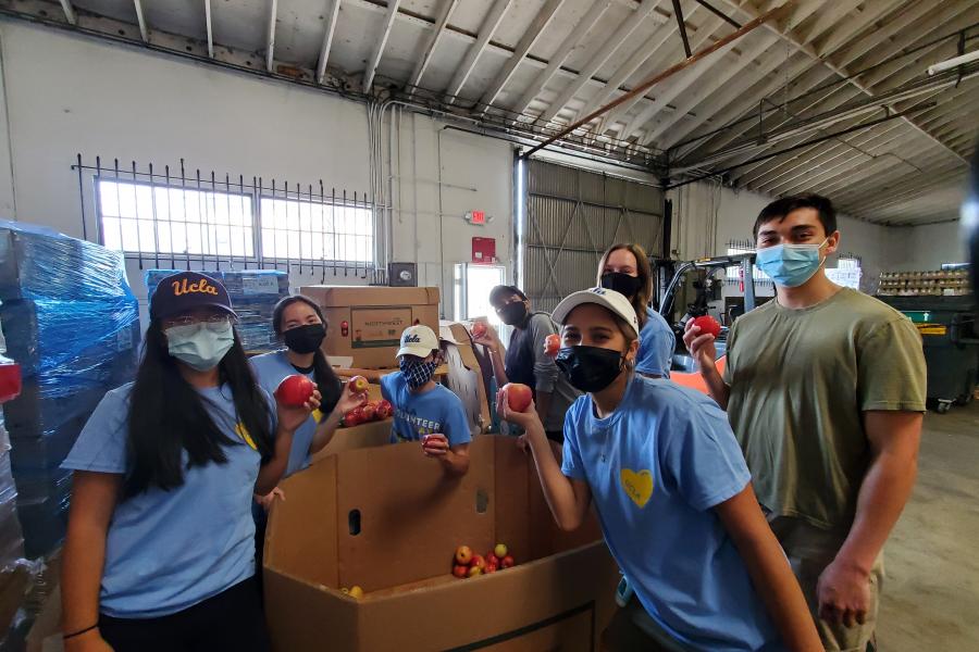 Seven masked volunteers pose around a cardboard crate, all holding apples and smiling. They are inside a warehouse and most of the volunteers are wearing a light blue UCLA Volunteer Day 2021 shirt.