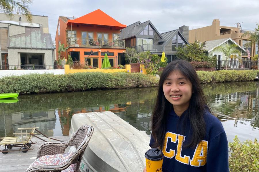 Photo of Alvina smiling in front of the Venice canals in Los Angeles. She is wearing a dark blue sweater with UCLA written in yellow lettering and holding a coffee cup from McDonalds.