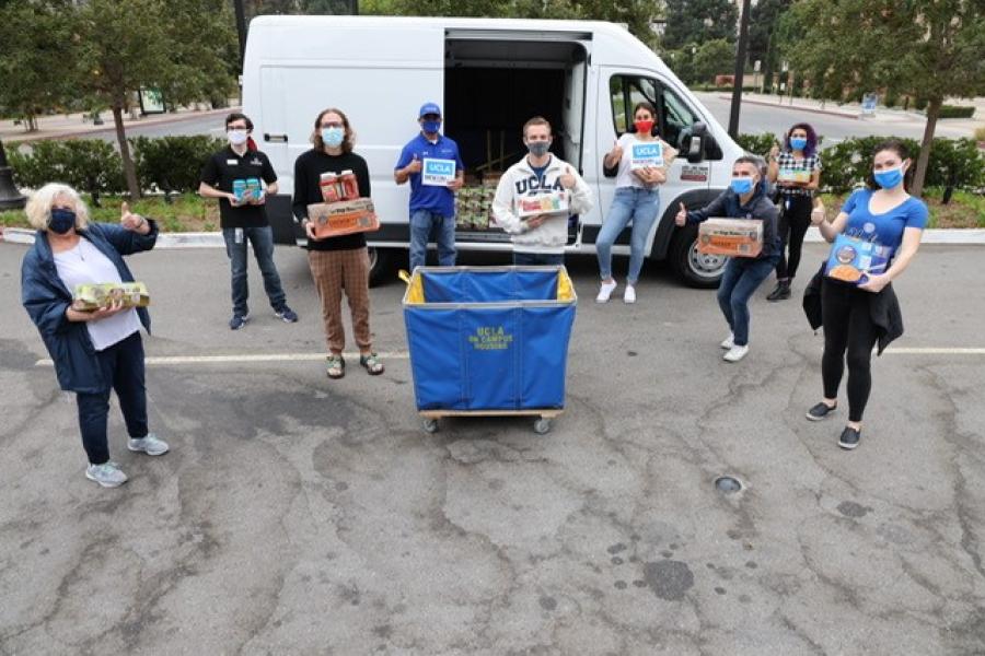 Nine volunteers stand around a UCLA Housing Cart. Each volunteer is masked and holding food and pantry items. They are standing in front of an open white catering van that is holding additional food.