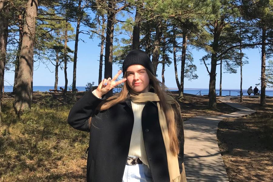 Photo of Nataly standing in front of some trees on a path, making a peace sign with her right hand. Nataly is wearing a black knit beanie, black coat, and tan scarf.